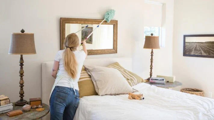 Woman dusting a mirror in a bright, neatly arranged bedroom with bed, pillows, lamps, and coastal decor