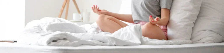 Person meditating cross-legged on a white bed with natural light and neutral bedroom decor